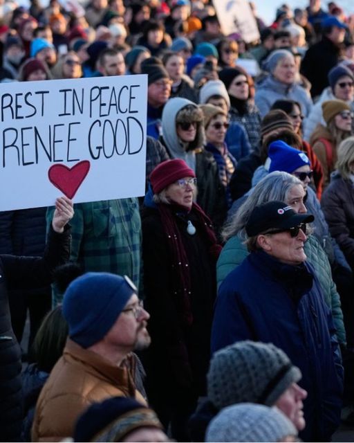Crowd at outdoor memorial holding REST IN PEACE RENEE GOOD sign with red heart